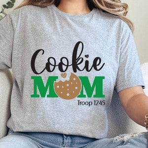 May include: Gray t-shirt with the text "Cookie MOM" in black and green. A brown cookie with a pink heart is in the middle of the word "MOM". The text "Troop 1245" is below the cookie.