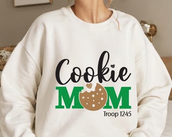 May include: White sweatshirt with the text "Cookie Mom" in black and green lettering. A brown cookie with a pink heart is in the middle of the word "Mom". The text "Troop 1245" is below the word "Mom".