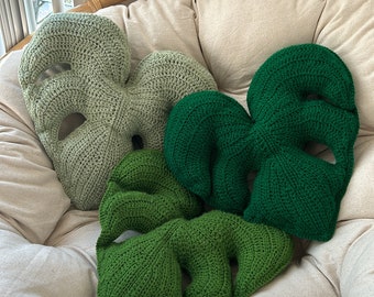May include: Three crocheted cushions shaped like monstera leaves. The cushions are green and light green. The cushions are sitting on a white chair.