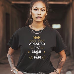 May include: Black t-shirt with the text "APLAUSO PA' MAMI Y PAPI" in gold and white. The design includes a crown, stars, and decorative flourishes. The shirt is worn by a person with tattoos and jewelry.