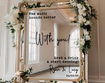 May include: A gold-framed mirror with white flowers draped around it. The mirror has a message written on it that says "The music sounds better with you! Grab a drink & start dancing! Ryan & Lucy 04/05/25"