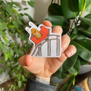 May include: A sticker featuring a cartoon Bitcoin coin riding a rollercoaster. The coin has a surprised expression and arms and legs. The rollercoaster is red and white and is ascending a hill.