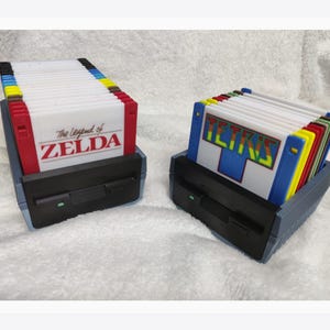 Puede incluir: Dos soportes de cartuchos de juego de estilo retro, cada uno con múltiples cartuchos de juego. Un soporte presenta "The Legend of Zelda" y el otro "Tetris". Los cartuchos son de varios colores y los soportes son negros y azules.