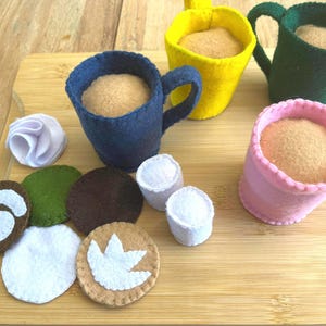 May include: A collection of felt play food items, including four colorful mugs filled with tan-colored felt, and various felt food pieces. The mugs are blue, yellow, green, and pink. Additional felt pieces include a swirl, a green circle, and two white marshmallow shapes.
