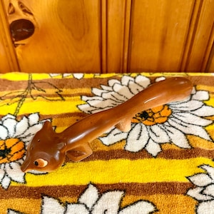 May include: A glossy brown ceramic squirrel figurine featuring a long, curved tail, small ears, and painted eyes. The squirrel is depicted in a horizontal, outstretched position with its head tilted downward, showcasing a smooth, reflective finish.