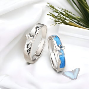 May include: Two silver rings with blue enamel. One ring features a lighthouse and the other features a sailboat. The rings are on a white surface with a blue butterfly.