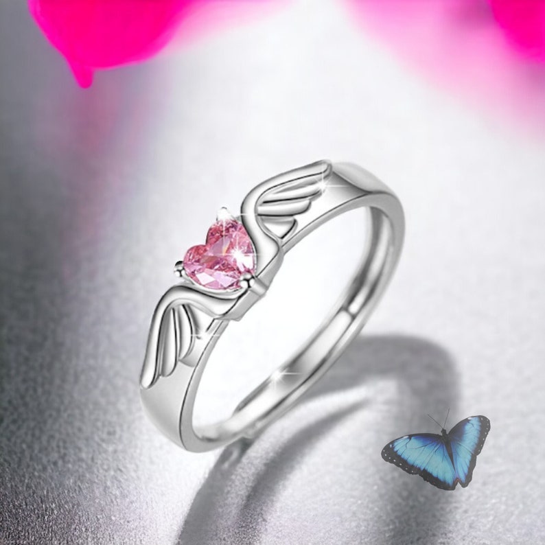 Puede incluir: Un anillo de plata con una piedra rosa en forma de coraz&oacute;n y dos alas a cada lado de la piedra.