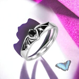 Puede incluir: Un anillo de plata con un dise&ntilde;o de murci&eacute;lago negro y una piedra preciosa negra. El anillo se coloca sobre un fondo rosa.