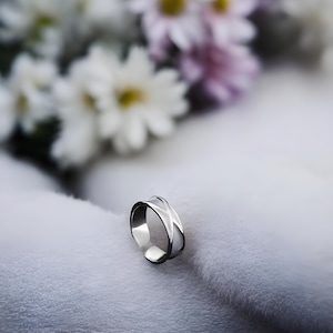 以下が含まれることがあります： クロスデザインのシルバーリング。リングは白いふわふわの表面の上に置かれています。