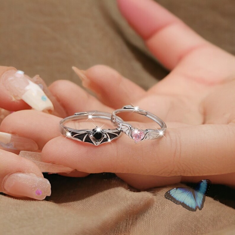 Puede incluir: Dos anillos de plata con piedras preciosas negras y rosas. El anillo de la izquierda presenta un dise&ntilde;o de murci&eacute;lago con una piedra preciosa negra. El anillo de la derecha presenta un dise&ntilde;o de coraz&oacute;n con una piedra preciosa rosa.