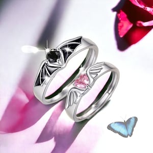Puede incluir: Dos anillos de plata con piedras en forma de coraz&oacute;n negras y rosas. El anillo de la izquierda tiene una piedra negra y un dise&ntilde;o de ala de murci&eacute;lago. El anillo de la derecha tiene una piedra rosa y un dise&ntilde;o de ala. Una mariposa azul est&aacute; en el fondo.