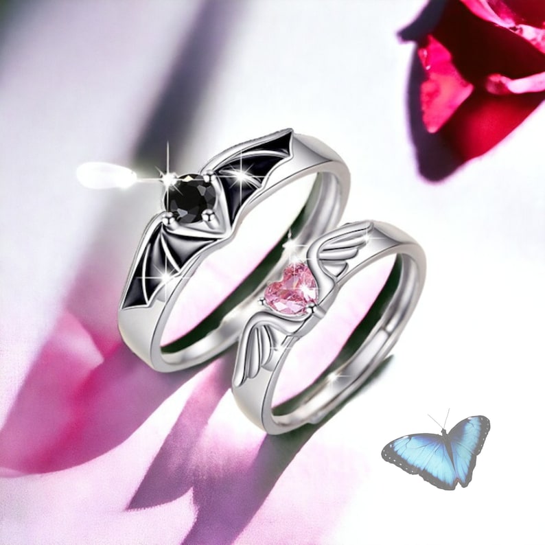 Puede incluir: Dos anillos de plata con piedras en forma de coraz&oacute;n negras y rosas. El anillo de la izquierda tiene una piedra negra y un dise&ntilde;o de ala de murci&eacute;lago. El anillo de la derecha tiene una piedra rosa y un dise&ntilde;o de ala. Una mariposa azul est&aacute; en el fondo.