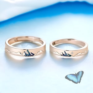 May include: Two gold rings with a mountain range design. One ring has a small diamond in the center of the mountain range. The rings are on a light blue background.