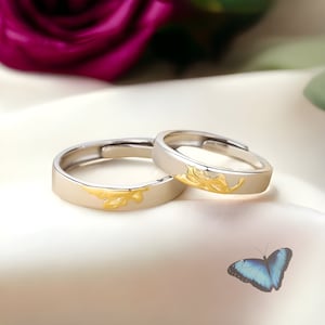 Puede incluir: Dos alianzas de plata con un diseño de hoja de oro. Las alianzas están sobre un fondo de satén blanco con una mariposa azul.