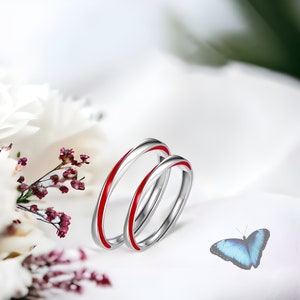 Può includere: Due fedi nuziali d'argento con una striscia rossa su ogni fede. Le fedi sono appoggiate su una superficie bianca con una farfalla blu sullo sfondo.