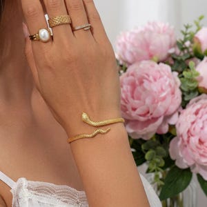 May include: A gold-toned snake-shaped cuff bracelet with a textured surface, worn alongside three gold-toned rings on the fingers. One ring features a central white oval pearl, another has a wide textured band, and the third is a thin band.