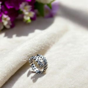 Puede incluir: Un anillo plateado con un diseño de hojas. El anillo está hecho de metal y presenta un patrón detallado de hojas alrededor de la banda. El anillo está sobre un fondo blanco con flores moradas en el fondo.
