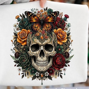 Puede incluir: Sudadera blanca con un diseño detallado de calavera rodeada de flores coloridas y una mariposa. La calavera está intrincadamente decorada con patrones florales, y las flores incluyen rosas y otras flores en tonos naranja, rojo y verde. La mariposa tiene alas naranjas y negras.