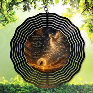 Op de afbeelding: Een ronde, gelaagde metalen windspinner met een hemels ontwerp. Het midden toont een engel en paardenbloem afbeelding met de tekst "Like Dandelion Seeds...Angel". Het buitenste frame is zwart met horizontale lijnen, hangend aan een ketting.