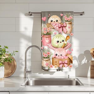 May include: A kitchen towel with a gray background features a whimsical design of cartoon dumplings, flowers, and hearts. The dumplings have smiling faces and are surrounded by pink and white accents. The towel is hanging on a silver bar above a kitchen sink.
