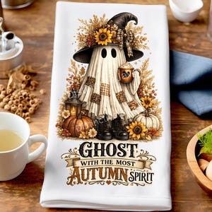 Peut inclure: Un torchon de cuisine blanc orné d'un fantôme portant un chapeau de sorcière, tenant une tasse et entouré de tournesols, de feuilles d'automne et de citrouilles. Sous l'illustration, le texte indique GHOST WITH THE MOST AUTUMN SPIRIT.