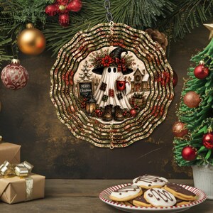 May include: A round, scalloped-edge metal wind spinner featuring a ghost wearing a witch hat, boots, and a plaid-patterned sheet, holding a red mug. The design includes a sign reading Merry Christmas, a lantern, and a snowy house background.