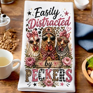 May include: A white kitchen towel featuring a graphic print of three chickens wearing sunglasses, bows, and hats. The text reads "Easily Distracted by PECKERS" in black and pink lettering. The design includes leopard print, pink flowers, and stars.