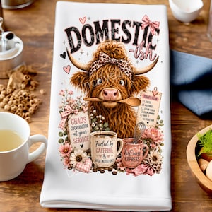 May include: A white kitchen towel featuring a brown Highland cow wearing a leopard print headband, holding a wooden spoon in its mouth. The design includes text reading DOMESTIC-ish, Chaos Coordinator, Fueled by Caffeine, and Hot Mess Express.