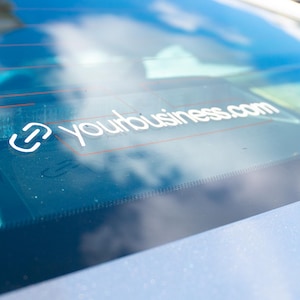 以下が含まれることがあります： 車の窓に貼られた白いステッカーで、"yourbusiness.com"のテキストと様式化されたロゴが表示されています。ステッカーは水色の車のリアウィンドウにあり、空が映っています。