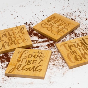 Puede incluir: Cuatro posavasos de madera con citas inspiradoras. Los posavasos presentan las frases "Salvado por la gracia", "Amar como Jesús", "Sé fuerte y valiente" y "Confía en el Señor".