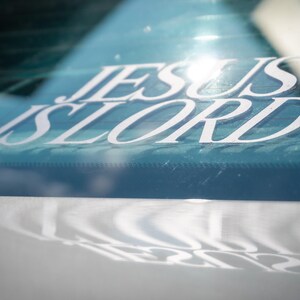May include: White vinyl decal with the words "JESUS IS LORD" in a stylized font, affixed to a car window. The decal is reflected on the car's surface below. The background is a light blue color.