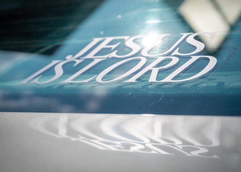 May include: White vinyl decal with the words "JESUS IS LORD" in a stylized font, affixed to a car window. The decal is reflected on the car's surface below. The background is a light blue color.