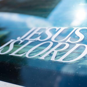 May include: White vinyl decal with the words "JESUS IS LORD" in a stylized font, applied to a car window. The background shows a blurred view of the sky and trees.