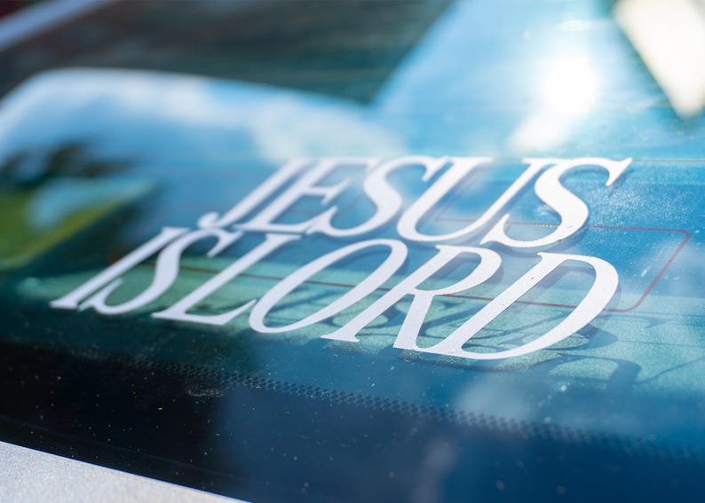 May include: White vinyl decal with the words "JESUS IS LORD" in a stylized font, applied to a car window. The background shows a blurred view of the sky and trees.
