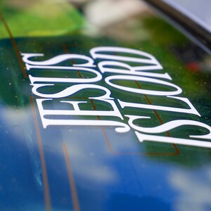 May include: White vinyl decal with the words "JESUS LORD" in a stylized font, applied to a car window. The background shows a blurred view of green and blue hues, suggesting an outdoor setting.