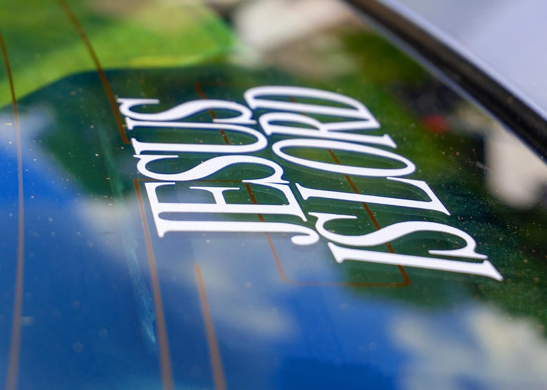May include: White vinyl decal with the words "JESUS LORD" in a stylized font, applied to a car window. The background shows a blurred view of green and blue hues, suggesting an outdoor setting.