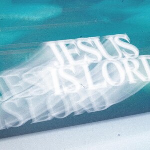 May include: An abstract image with the words "JESUS IS LORD" in white, layered over a blurred, teal and blue background. The text appears to be slightly transparent, creating a ghostly effect.