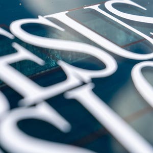 May include: Close-up of a blue surface with the word "JESUS" in large, white, stylized letters. The letters are slightly blurred, creating a soft focus effect. The background is a deep blue color.