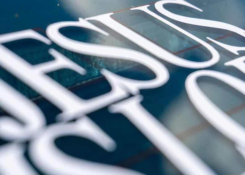May include: Close-up of a blue surface with the word "JESUS" in large, white, stylized letters. The letters are slightly blurred, creating a soft focus effect. The background is a deep blue color.