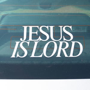 May include: White vinyl decal with the words "JESUS IS LORD" in a bold, serif font. The decal is affixed to the rear window of a vehicle, with the text prominently displayed against the dark background.