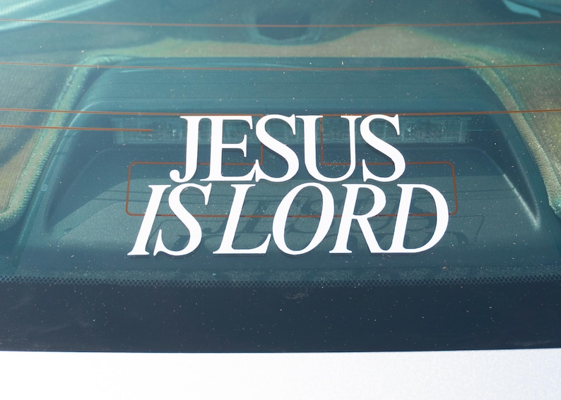 May include: White vinyl decal with the words "JESUS IS LORD" in a bold, serif font. The decal is affixed to the rear window of a vehicle, with the text prominently displayed against the dark background.