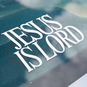 May include: White vinyl decal with the words "JESUS IS LORD" in a bold, serif font. The decal is applied to a car's rear window, with a green-tinted background and visible defroster lines.
