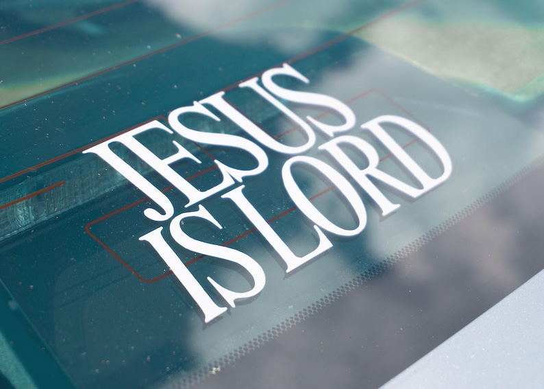 May include: White vinyl decal with the words "JESUS IS LORD" in a bold, serif font. The decal is applied to a car's rear window, with a green-tinted background and visible defroster lines.