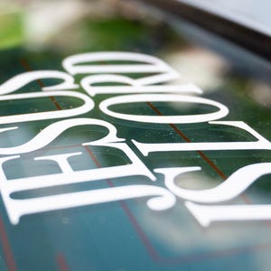 May include: White decal on a car window. The decal reads "JESUS LORD" in a stylized, serif font. The background is a blurred view of greenery.