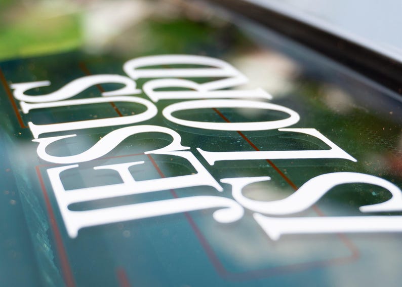 May include: White decal on a car window. The decal reads "JESUS LORD" in a stylized, serif font. The background is a blurred view of greenery.