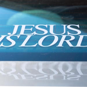 May include: A white decal on a blue surface reads "JESUS IS LORD" in a bold, sans-serif font. The decal is affixed to a window, with a reflection of the text visible below. The background is a blurred view of the outside.