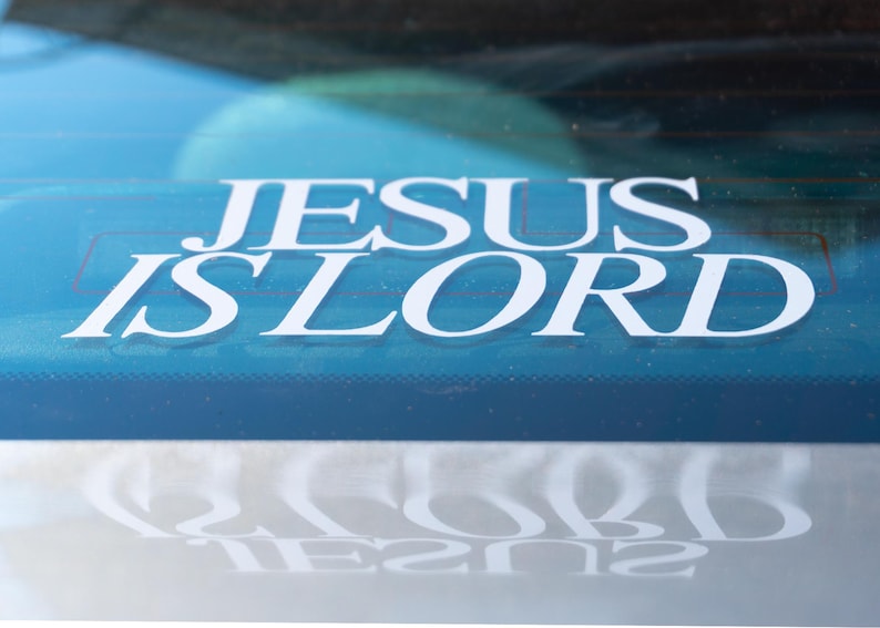 May include: A white decal on a blue surface reads "JESUS IS LORD" in a bold, sans-serif font. The decal is affixed to a window, with a reflection of the text visible below. The background is a blurred view of the outside.