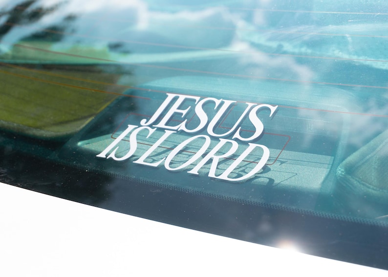 May include: White vinyl decal with the words "JESUS IS LORD" applied to a car window. The decal is in a stylized font and is visible against the background of the car's interior and the sky.