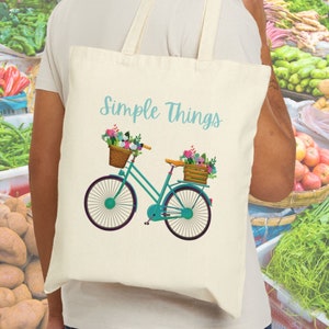 May include: Cream-colored tote bag with the text "Simple Things" in light blue. The bag features a teal bicycle illustration with flower baskets. The bicycle has brown seats and black wheels. The background is a farmer's market.