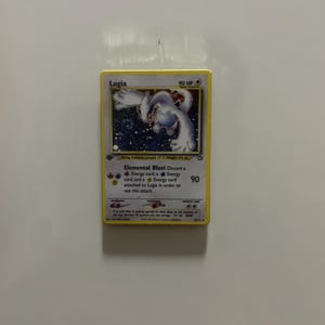 May include: A Lugia trading card featuring a white creature illustration, 90 HP, and the Elemental Blast attack text. The card displays fire, water, and lightning energy symbols, with a yellow border and detailed game statistics printed below.